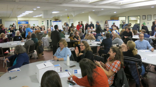 Photo of hundreds of people sitting around many round tables in a senior center meeting room.